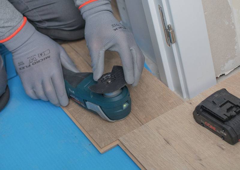 Ein Arbeiter schneidet Laminatboden mit einem Multifunktionswerkzeug in der Nähe einer Tür. Schutzhandschuhe und Akkupack sind auf der blauen Unterlage sichtbar.