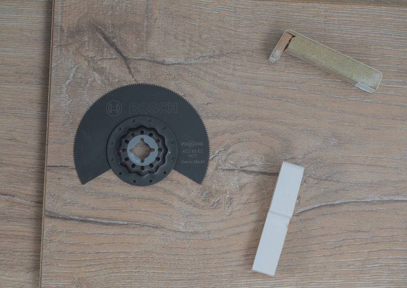 Ein Bosch-Sägeblatt für Oszillationssägen auf einem Holzboden neben zwei kleinen rechteckigen Teilen, eines weiß und eines beige.