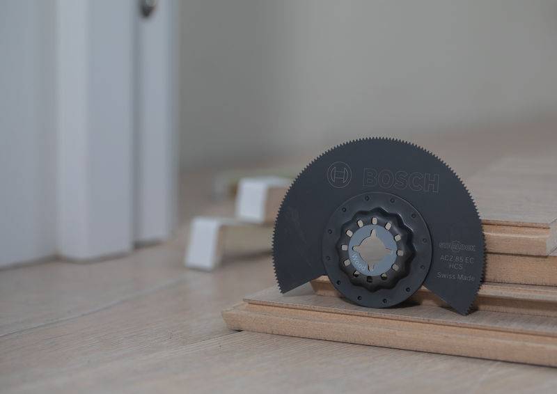 Ein Multitool-Kreissägeblatt ruht auf gestapelten Holzbrettern in der Nähe einer Wand und deutet auf eine Holzbearbeitungs- oder Hausrenovierungsprojekt-Anordnung hin.