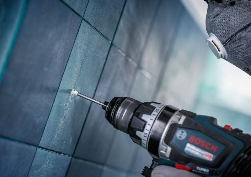 Eine Person benutzt einen Bosch-Elektrobohrer, um ein Loch in eine geflieste Wand zu bohren, und trägt dabei Schutzhandschuhe.