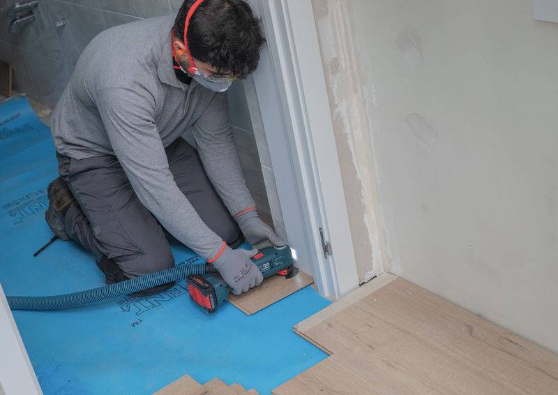 Eine Person verlegt Holzboden mit einem Elektrowerkzeug, trägt eine Schutzbrille und einen Gehörschutz. Blaue Unterlagematerialien sind darunter sichtbar.