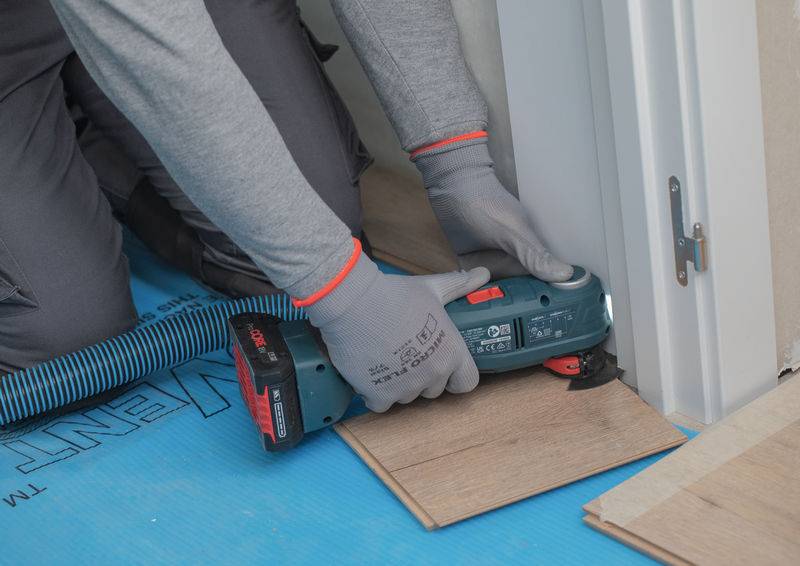 Arbeiter schneidet Laminatboden mit einer elektrischen Säge zurecht, wobei er ihn an einen weißen Türrahmen anpasst, während der Installation.