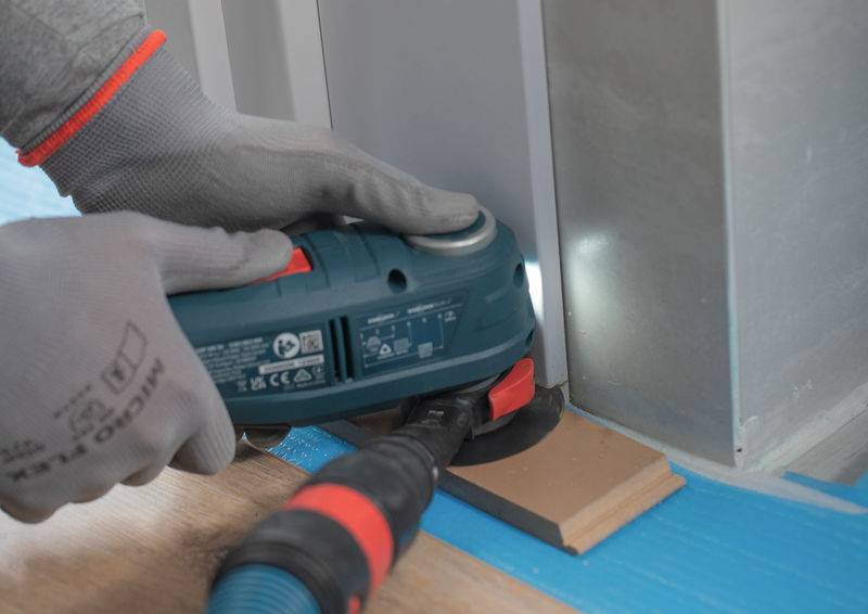 Eine Person verwendet ein Elektrowerkzeug, um ein Stück Holzfußboden neben einer Wand zu trimmen und trägt dabei Schutzhandschuhe.