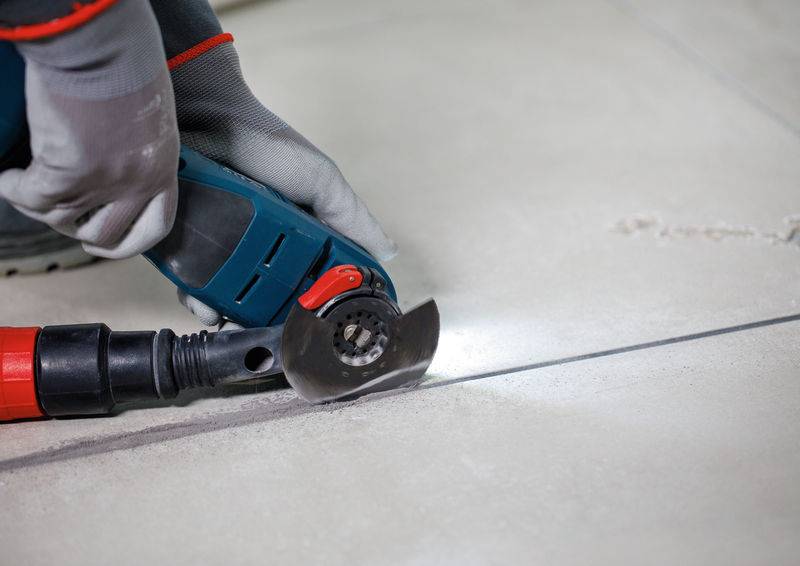 Eine Person in Schutzhandschuhen verwendet ein elektrisches Schneidewerkzeug, um eine gerade Linie auf einer großen Fliese zu ritzen, wobei Staub entsteht.