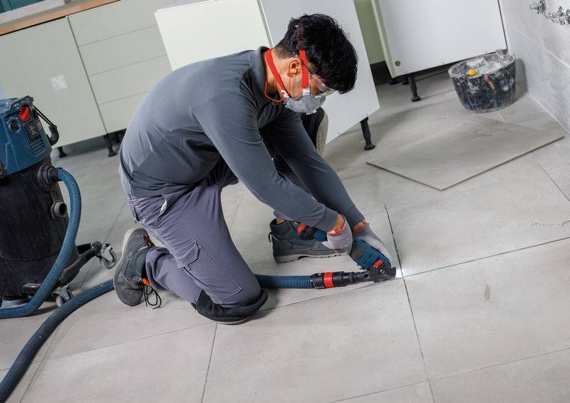 Eine Person in Schutzausrüstung verwendet ein Werkzeug, um Bodenfliesen in einem Renovierungsprojekt zu schneiden.