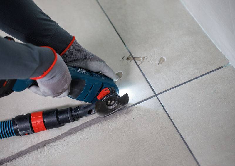 Eine Person, die ein Elektrowerkzeug zum Schneiden von Fliesen entlang der Fugenlinie verwendet, trägt Schutzhandschuhe und stellt eine präzise Ausrichtung mit den Fliesenfugen sicher.