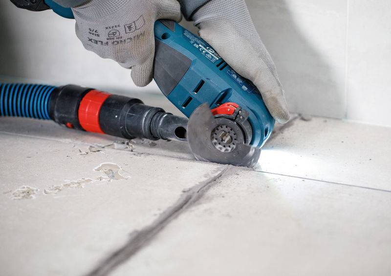 Eine Person benutzt eine Elektrosäge, um Fliesen auf einem Fußboden zu schneiden, wobei Staub von einem angeschlossenen Schlauch abgesaugt wird, um Sauberkeit und Präzision zu gewährleisten.