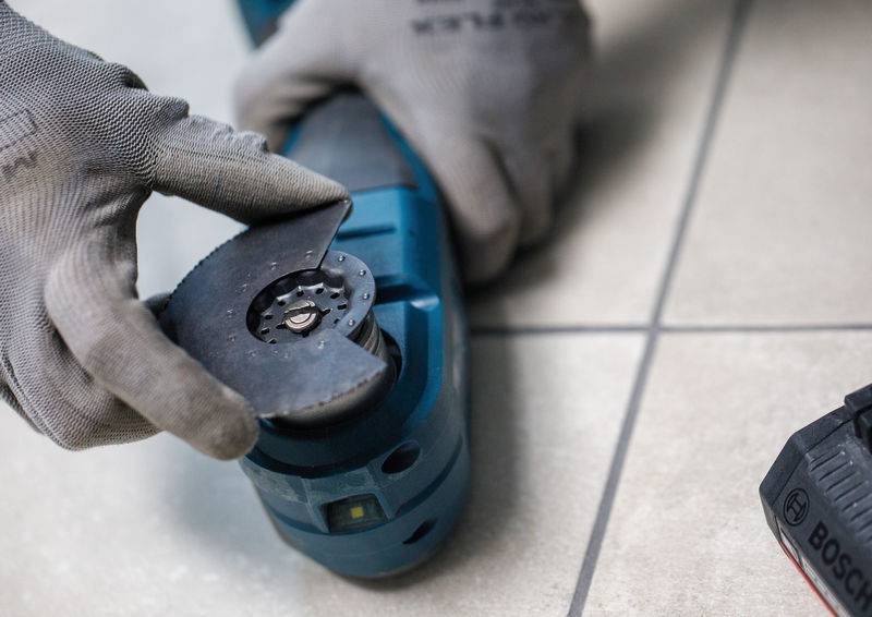 Eine Person mit Handschuhen montiert oder stellt ein Elektrowerkzeug auf einer gefliesten Oberfläche ein. Die Hände positionieren einen kreisförmigen Aufsatz.