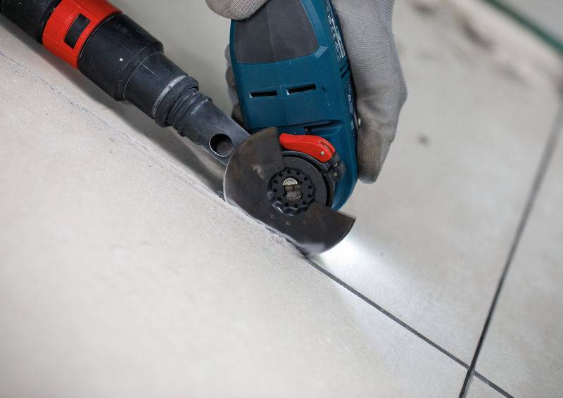 Eine Hand in einem Handschuh bedient ein Elektrowerkzeug zum Schneiden von Fugenmaterial zwischen Bodenfliesen mit Fokus auf Präzision und Sicherheit.