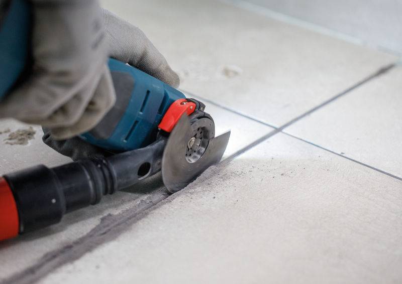 Eine Person mit Handschuhen nutzt ein Elektrowerkzeug, um entlang einer markierten Linie beigefarbene Bodenfliesen durchzuschneiden, wobei Staub um den Schnitt sichtbar ist.