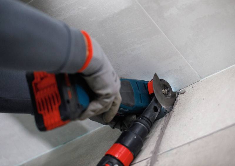 Eine Person nutzt ein Elektrowerkzeug, um graue Fliesen in einer Ecke zu schneiden, und trägt dabei Handschuhe zum Schutz.