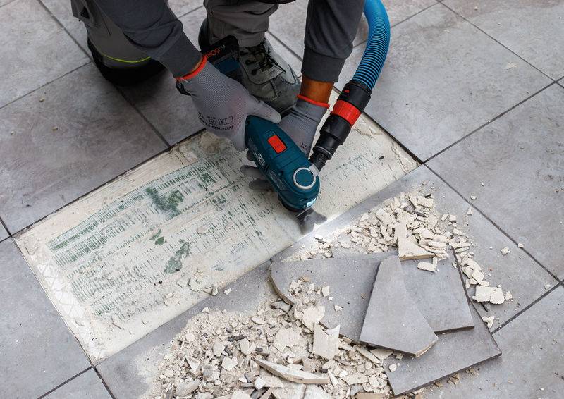 Eine Person benutzt ein Elektrowerkzeug, um Fugenmaterial zwischen Bodenfliesen zu entfernen; abgesplitterte Fliesen und Schutt sind in der Nähe verstreut.