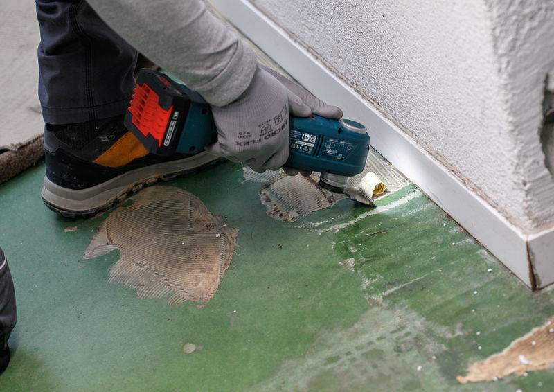 Eine Person nutzt ein Elektrowerkzeug, um Klebstoff von einem grünen Boden in der Nähe einer weißen Wand zu entfernen. Die Person trägt Handschuhe und Schutzschuhe.