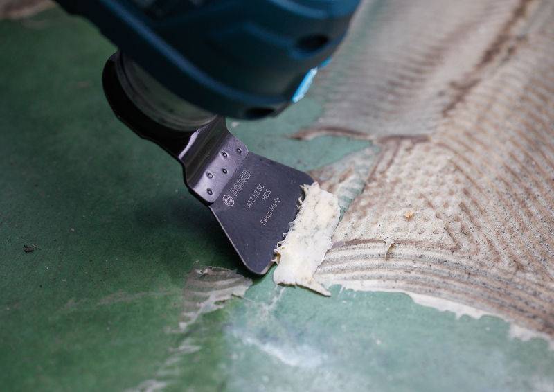 Ein Elektrowerkzeug mit einer Schabeklinge entfernt Klebstoff von einer grünen Oberfläche und bereitet sie für Reparaturen oder Installationen vor.