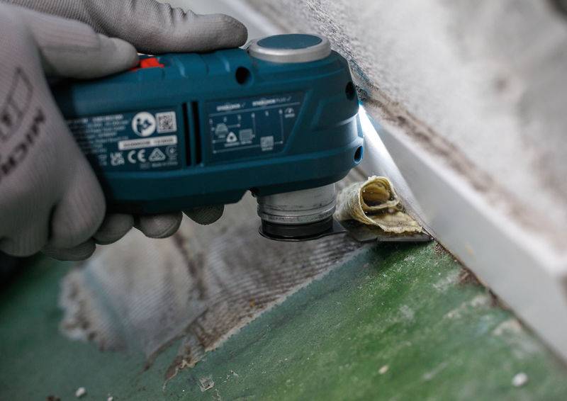 Eine Person benutzt ein elektrisches Handwerkzeug, um Bodenbeläge in der Nähe einer weißen Wand zu schneiden, trägt Schutzhandschuhe. Ein Materialstück wird entfernt.