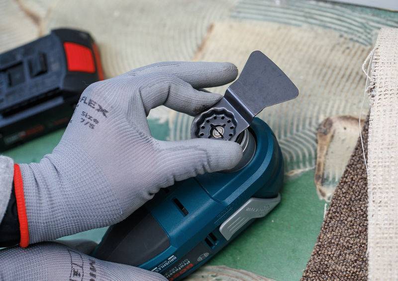 Eine Person mit Handschuhen befestigt eine Klinge an einem Elektrowerkzeug und bereitet es für die Verwendung vor. Das Werkzeug wird auf einer Arbeitsfläche platziert.