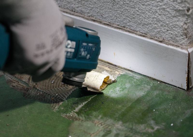 Eine Person verwendet ein Elektrowerkzeug, um Klebstoff von einem grünen Boden neben einer Fußleiste zu entfernen und legt dabei eine zerkratzte, unebene Oberfläche frei.