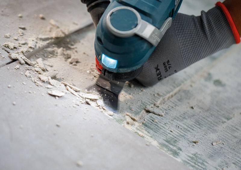 Eine Person mit einem Handschuh benutzt ein Elektrowerkzeug, um Klebstoff von einem Betonboden abzukratzen, entfernt Verschmutzungen und glättet die Oberfläche.