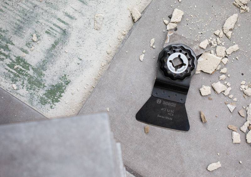 Oszillierwerkzeug-Sägeblatt auf Betonboden inmitten von Schutt, in der Nähe einer grün gestrichenen Fläche. Blatt gekennzeichnet: BiM ACZ 85 EB.