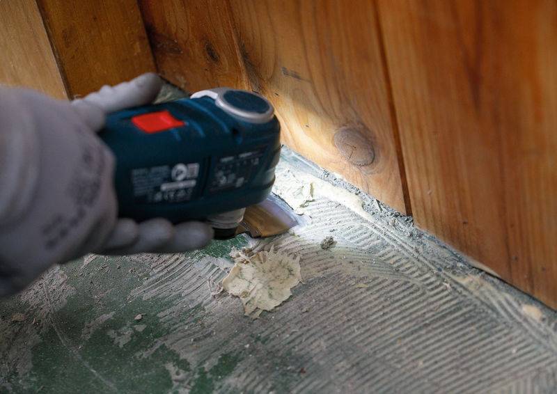 Eine Person benutzt ein elektrisches Werkzeug, um Klebereste von einem Betonboden neben einer Holzwand zu entfernen, was auf Renovierungsarbeiten hinweist.