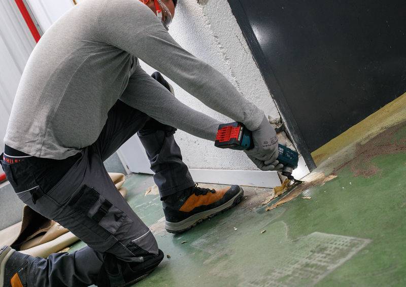Eine Person kniet auf einem Betonboden und verwendet ein Elektrowerkzeug, um in die Basis einer Wand zu schneiden. Sie trägt Schutzausrüstung und Arbeitsschuhe.