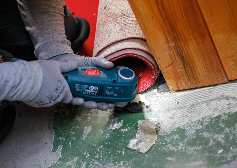 Eine Person benutzt ein Elektrowerkzeug zum Schneiden von Teppich in der Nähe einer Holzoberfläche, wobei sie Handschuhe trägt, um die Sicherheit zu gewährleisten. Dies unterstreicht eine Renovierungsaktivität.