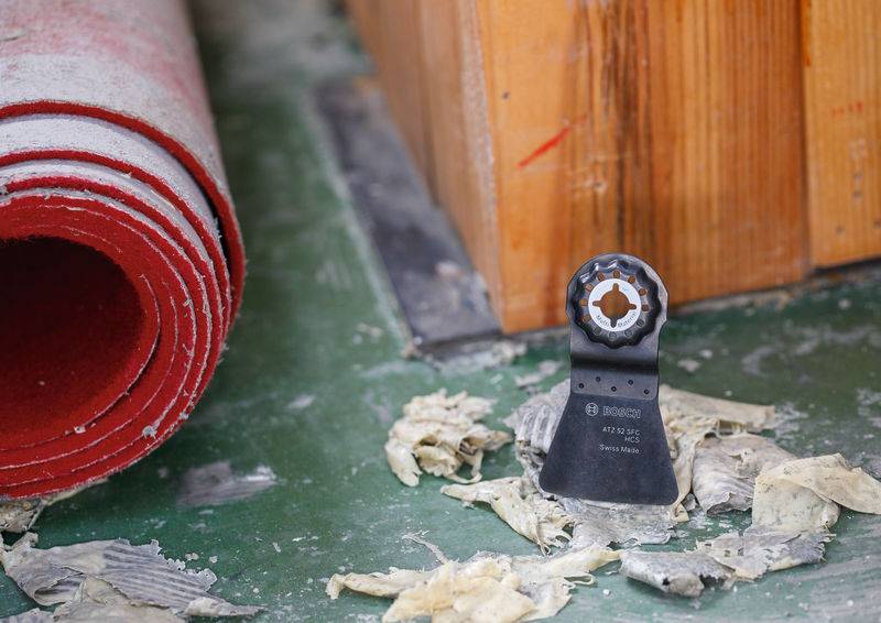 Ein zerlegter roter Teppichroller liegt auf einem grünen Boden neben einem Holzobjekt. Eine Sägeblatt steht aufrecht zwischen verstreutem Schutt in der Nähe.