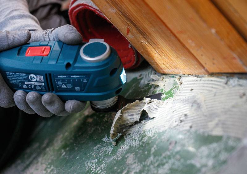 Eine Person mit Handschuhen verwendet ein blaues Elektrowerkzeug mit Licht, um Klebstoff von einer grünen Oberfläche zu entfernen, in der Nähe einer Holzkonstruktion.