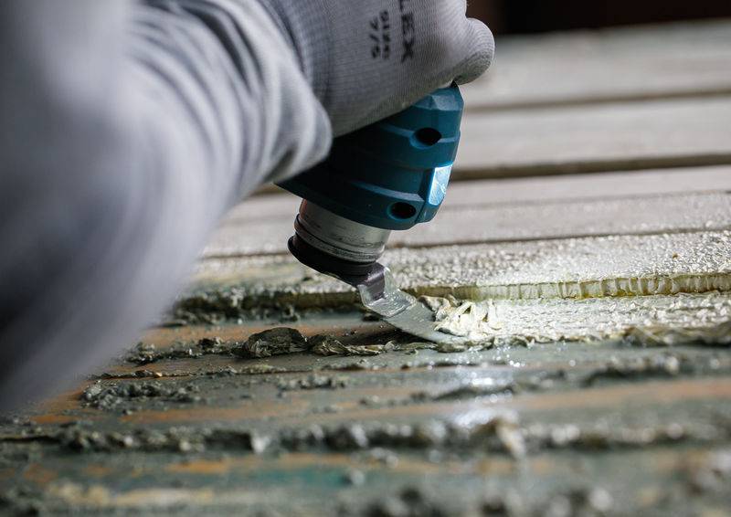 Eine Person mit Handschuhen benutzt ein Elektrowerkzeug, um Farbe von einer Holzoberfläche abzukratzen und konzentriert sich darauf, Schichten zu entfernen, um das bloße Holz freizulegen.