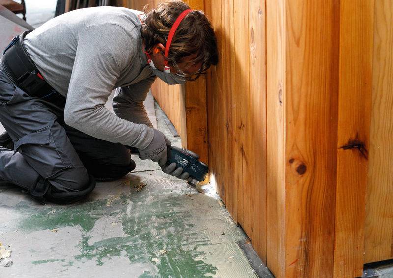 Eine Person in Schutzausrüstung schleimt einen hölzernen Zaun im Inneren und konzentriert sich darauf, seine Oberfläche zu glätten. Staub und Schleifspäne sind auf dem Boden sichtbar.