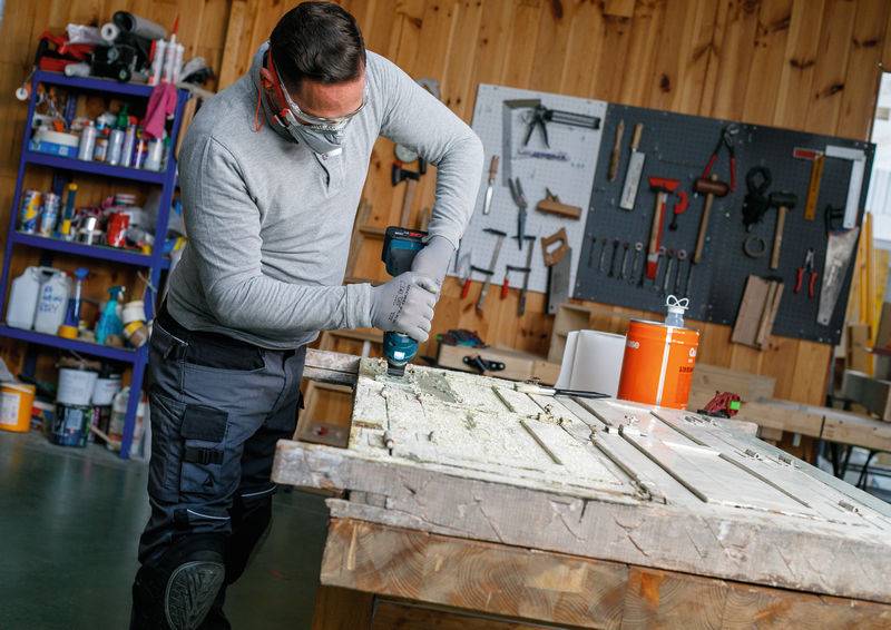 Eine Person in Schutzausrüstung benutzt einen Elektroschrauber an einer Holztür in einer Werkstatt, umgeben von Werkzeugen und Regalen mit Materialien.