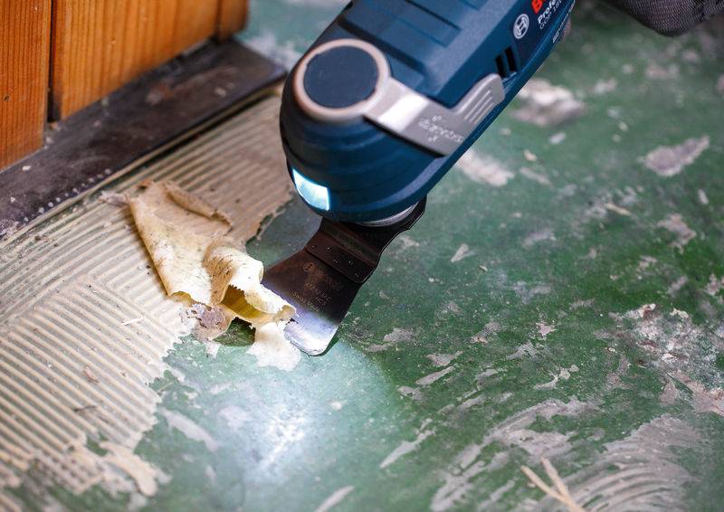 Ein blaues Elektrowerkzeug mit flacher Klinge entfernt Klebstoff von einem grünen Boden, in der Nähe einer Holzoberfläche, was einen Renovierungsprozess zeigt.