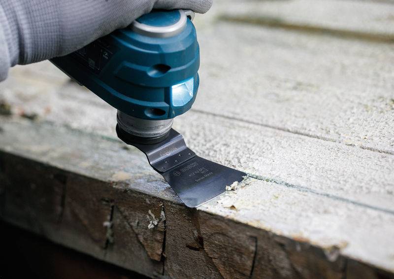 Eine Person verwendet ein blaues oszillierendes Multifunktionswerkzeug mit einer flachen Klinge, um Farbe von einer Holzoberfläche abzukratzen, während sie einen grauen Handschuh trägt.