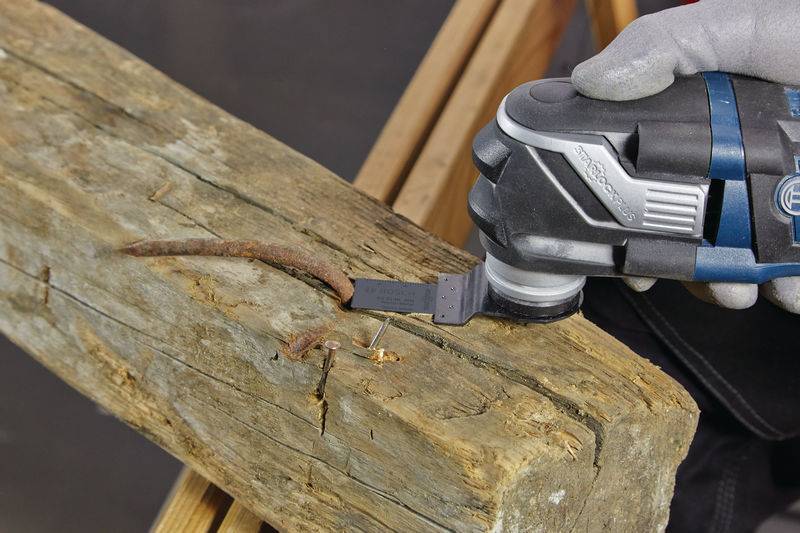Die behandschuhte Hand einer Person benutzt ein oszillierendes Multitool, um einen rostigen Nagel zu schneiden, der aus einem verwitterten Holzbalken hervorragt.