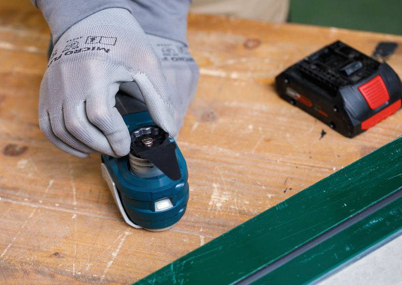 Eine Person mit grauen Handschuhen befestigt eine schwarze Schneideklinge an einem blauen Elektrowerkzeug auf einer Holzwerkbank.