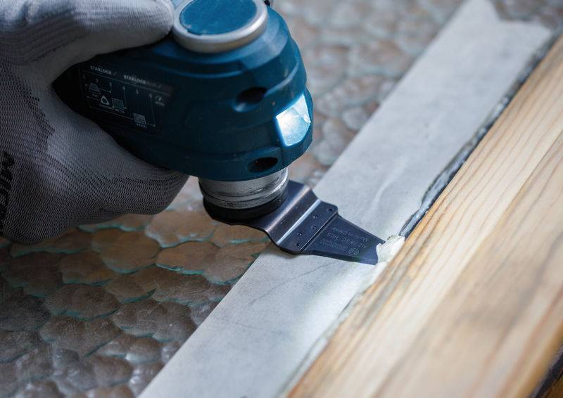 Eine behandschuhte Hand benutzt ein blaues Elektrowerkzeug mit einer flachen Klinge, um weiße Farbe von einer Holzoberfläche zu entfernen und enthüllt dabei eine wabenförmige Glastextur.