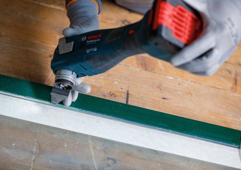 Eine Person, die ein Elektrowerkzeug mit einer Metallschabeaufsatz auf einer Holzoberfläche verwendet, trägt Handschuhe zur Sicherheit und konzentriert sich auf präzise Arbeit.