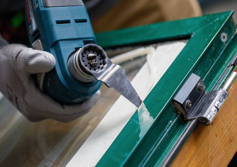 Nahaufnahme einer Person, die ein oszillierendes Multiwerkzeug verwendet, um alte Farbe von einem grünen Holzrahmen mit einem Metallscharnier zu entfernen.