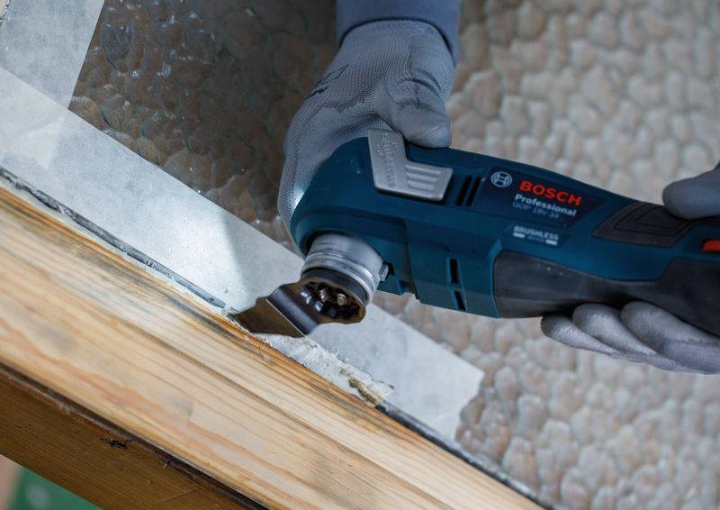 Eine Person, die Handschuhe trägt, verwendet ein Bosch Professional Elektrowerkzeug, um Farbe von einer Holzoberfläche zu entfernen, wobei Schutzband entlang der Kanten angebracht ist.