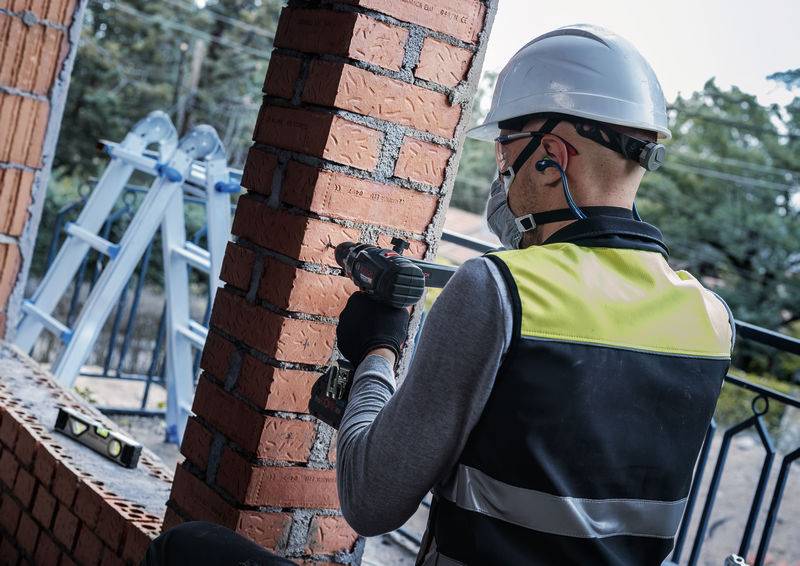 Ein Bauarbeiter in Schutzausrüstung bearbeitet mit einem Werkzeug eine Backsteinmauer. Eine Leiter und Bäume sind im Hintergrund sichtbar.