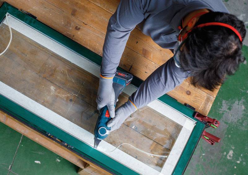 Eine Person mit Handschuhen verwendet ein Elektrowerkzeug, um einen Fensterrahmen auf einem Holztisch in einer Werkstattumgebung durchzuschneiden.
