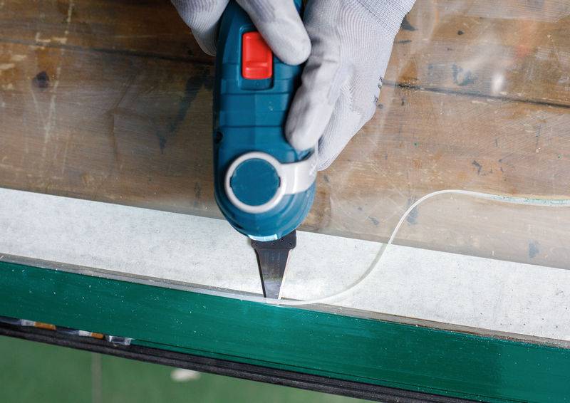 Eine Person mit Handschuhen verwendet ein blaues Universalmesser, um weiße Zierleisten an einer Glasscheibe zu schneiden, was Präzision beim Handwerk oder bei der Montage hervorhebt.