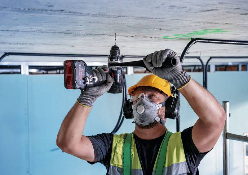 Ein Bauarbeiter in Schutzausrüstung, einschließlich Helm und Maske, benutzt einen Bohrer an der Decke. Es werden Schutzbrille, Gehörschutz und Handschuhe getragen.