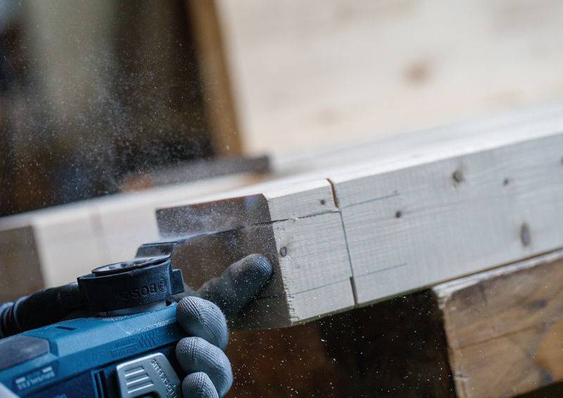 Eine Nahaufnahme einer behandschuhten Hand, die ein Elektrowerkzeug verwendet, um ein Stück helleres Holz zu schneiden, mit Sägespänen in der Luft.