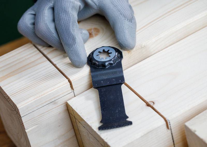 Eine behandschuhte Hand hält ein Stahlmulti-Funktionswerkzeug-Blatt gegen eine Holzoberfläche mit präzisen Schnitten, was auf Holzbearbeitung oder Zimmermannsarbeit hinweist.