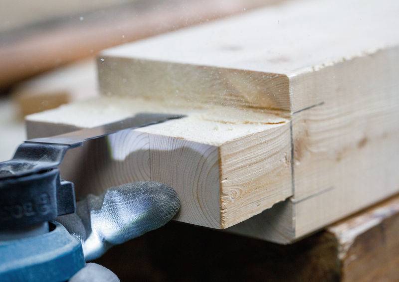 Eine Nahaufnahme einer Säge, die in einen Holzbalken schneidet, mit sichtbaren Holzspänen. Der Balken ist sicher in einer Werkbank eingespannt.