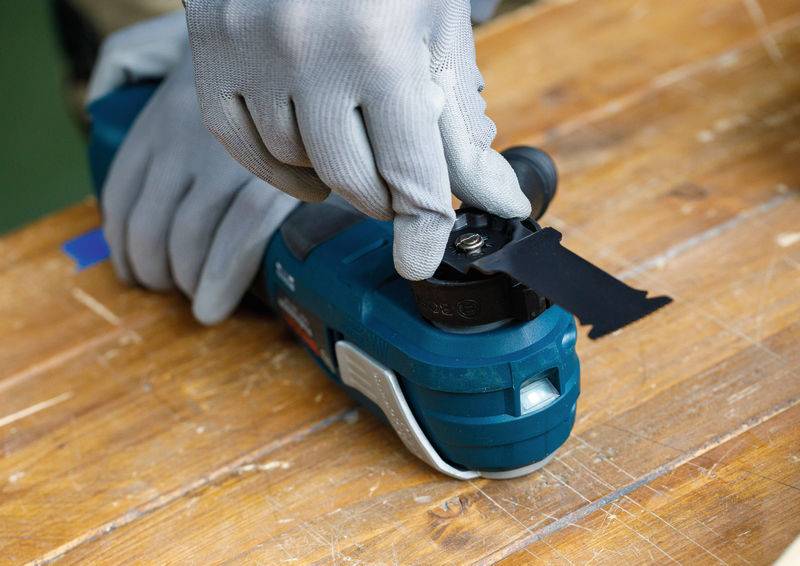Eine Person mit Handschuhen stellt die Klinge eines blauen Elektrowerkzeugs auf einer Holzoberfläche ein und demonstriert dabei Wartung oder Werkzeugvorbereitung.