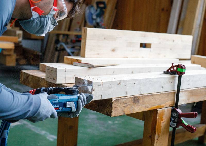 Eine Person mit Schutzbrille benutzt ein Elektrowerkzeug, um Holzbretter auf einer Werkbank in einer Werkstatt zu schneiden.