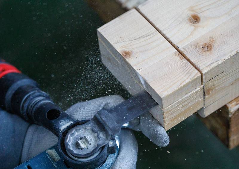 Eine Person verwendet ein Elektrowerkzeug, um ein Stück helles Holz zu schneiden. Sägespäne sind sichtbar, während das Werkzeug durch das Holz schneidet.
