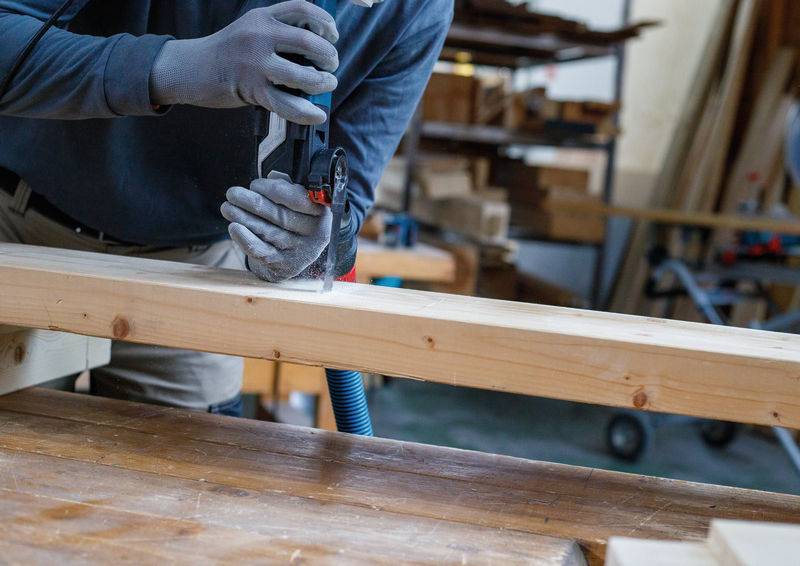 Eine Person benutzt ein Elektrowerkzeug, um in ein Holzbrett in einer Schreinerwerkstatt zu bohren, mit Holzmaterialien und Werkzeugen, die im Hintergrund sichtbar sind.